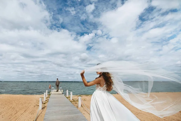 Junge Braut, modische und schöne brünette Modell Mädchen in stilvollem Hochzeitskleid mit einem Schleier, der im Wind flattert, geht zu dem Bräutigam, der am Pier wartet — Stockfoto