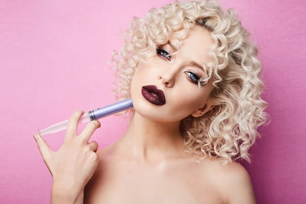 Schöne und modische Model-Mädchen mit erstaunlichen blauen Augen, mit lockigen blonden Haaren und mit professionellem hellen Make-up, macht Injektion selbst mit einer blauen Spritze und posiert vor dem rosa Hintergrund — Stockfoto