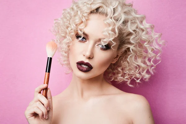 Fashionabla och eleganta modell flicka med blå ögon och lockigt blont hår, med professionell ljus makeup poserar med makeup borste i hennes hand på studio, isolerad på rosa bakgrund — Stockfoto