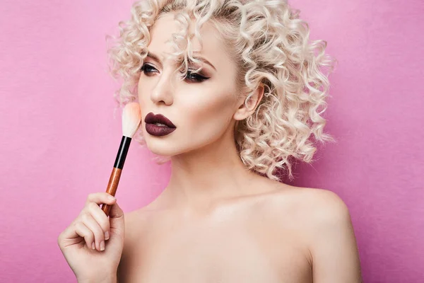 Fashionabla och eleganta modell flicka med blå ögon och lockigt blont hår, med professionell ljus makeup poserar med makeup borste i hennes hand på studio, isolerad på rosa bakgrund — Stockfoto