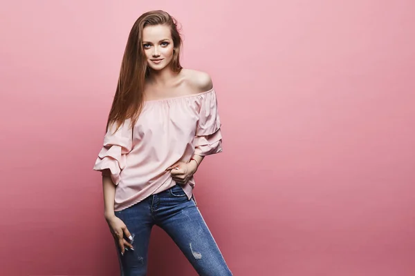 Hermosa y de moda chica modelo rubio con maquillaje brillante profesional en blusa de satén de moda y en vaqueros azules posando en el fondo rosa — Foto de Stock
