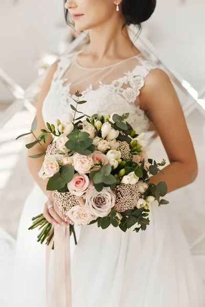 Menina modelo morena bonita e elegante em um vestido de noiva elegante branco com um grande e luxuoso buquê de flores exóticas em suas mãos, posando no interior — Fotografia de Stock