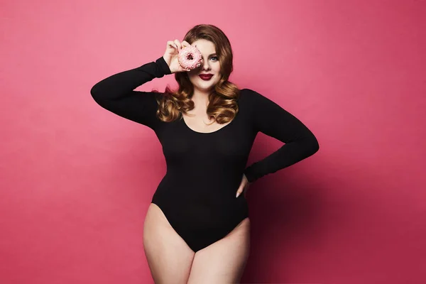 Plus size sexy model girl, fashionable blonde in black bodysuit, with bright makeup and with donut in her hand smiling and posing at pink background in studio — Stock Photo, Image