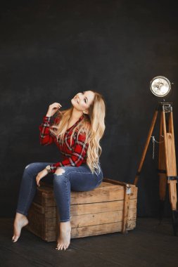 Boyutu seksi model kız, karanlık iç poz ahşap göğüs mutlu sarışın parlak makyaj ile ve kırmızı kareli gömlek ve şık bir kot pantolon güzel bir gülümseme ile oturuyor