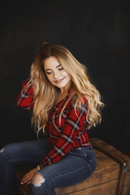 Plus size sexy model girl, fashionable blonde with bright makeup and with a beautiful smile in red plaid shirt and in a stylish jeans sits on the wooden chest posing at dark interior clipart