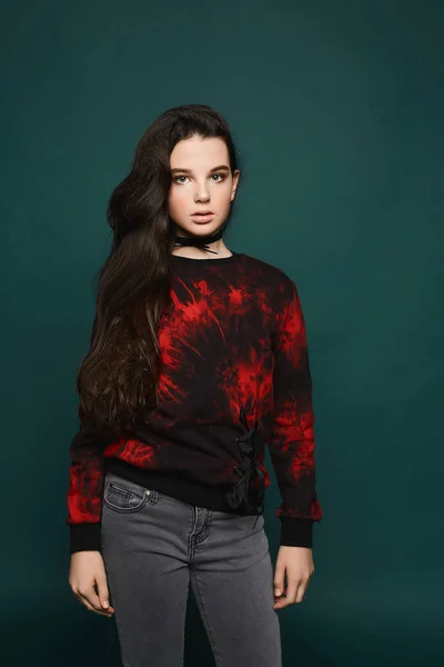Beaybeautiful und modische brünette Teenager-Modell Mädchen in stilvollen grauen Jeans und im schwarzen Sweatshirt mit rotem Muster posiert vor dunkelgrünem Hintergrund — Stockfoto