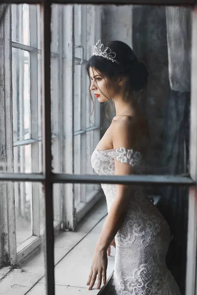 Menina modelo morena sexy e bonita em vestido branco de renda com ombros nus com diadema na cabeça fica perto da janela e olhando para fora esperando por seus homens a foto é feita através de uma janela — Fotografia de Stock