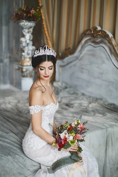 Beautiful fashionable bride, young model brunette woman in stylish wedding dress with naked shoulders with bouquet of flowers in her hands sits on the bed and posing in luxury vintage interior