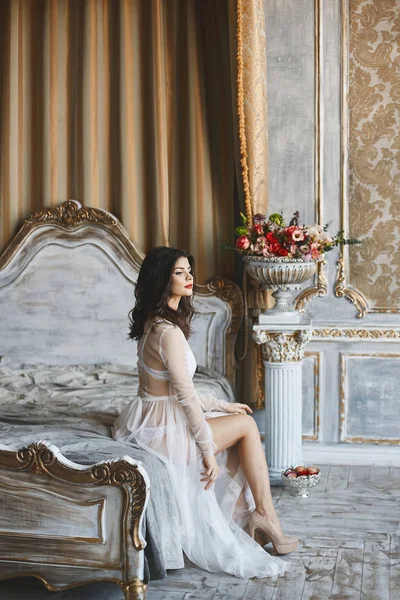 Sexy e bella bruna, alla moda giovane donna modello con corpo perfetto in lingerie bianca e in peignoir elegante si siede su un letto — Foto Stock