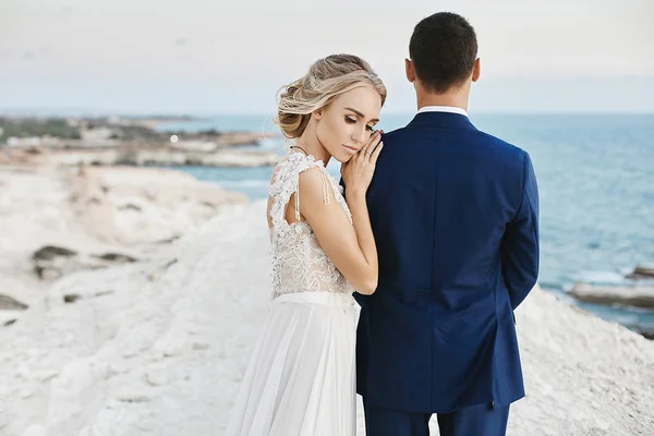 美丽的年轻金发模特女孩在时尚的白色蕾丝礼服靠在英俊的男人在时尚的蓝色西装和在亚得里亚海沿岸的白色岩石上摆姿势 — 图库照片