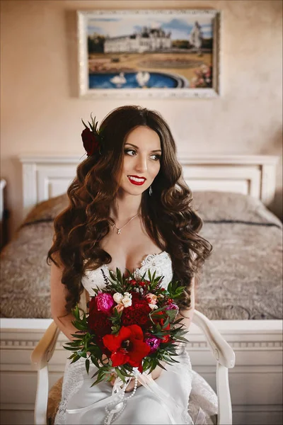 Menina modelo morena elegante e bonita com maquiagem brilhante e com um sorriso brilhante, em um vestido de noiva branco e com um grande buquê de luxo de flores exóticas em suas mãos posando no interior — Fotografia de Stock