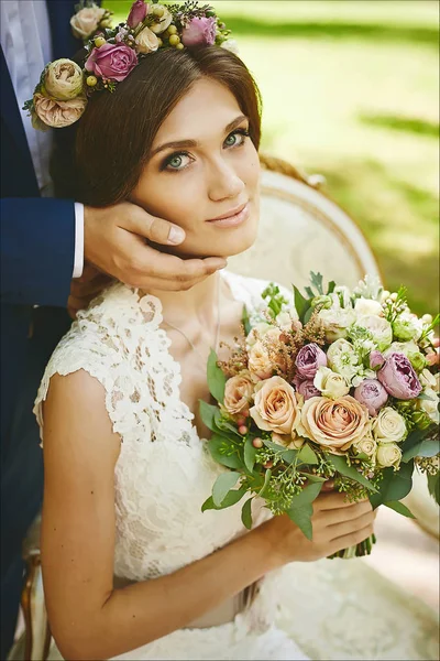 Modische und schöne brünette Modell Mädchen mit stilvoller Hochzeitsfrisur und mit hellem Make-up in trendigen Spitzenkleid sitzt auf einem Stuhl mit dem Strauß exotischer Blumen in ihren Händen — Stockfoto