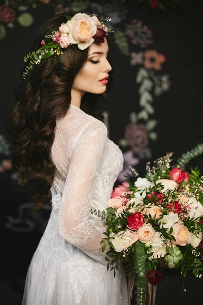 Menina modelo morena elegante e bonita com maquiagem brilhante e com uma coroa de flores na cabeça em um vestido de renda elegante e com um grande buquê de luxo de flores em suas mãos posando no interior — Fotografia de Stock