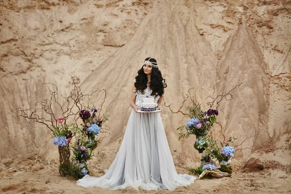 Menina modelo morena bonita e elegante com jóias na cabeça no vestido de renda elegante com o bolo de casamento criativo e elegante em suas mãos posando em um deserto — Fotografia de Stock