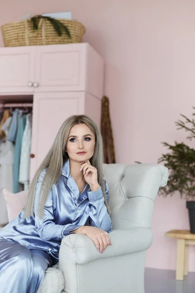 Menina modelo loira encantadora e elegante com olhos azuis em pijama de cetim elegante senta-se na poltrona vintage e posando no interior rosa claro — Fotografia de Stock