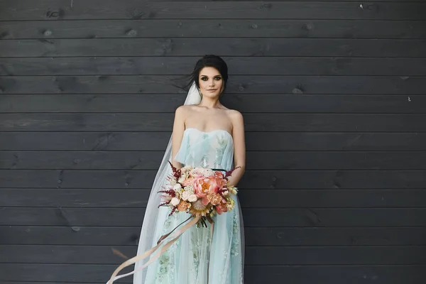 Sexy und schöne brünette Modell Mädchen mit hellem Make-up im modischen Hochzeitskleid und im Schleier mit dem großen Luxus-Blumenstrauß in ihren Händen posiert vor der dunklen Holzwand — Stockfoto