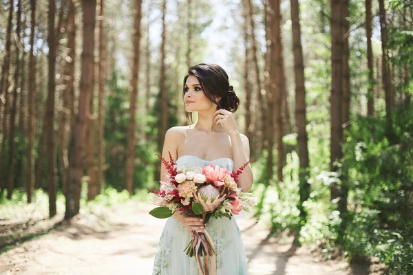 Joyeux et souriant brunette fille sexy avec un maquillage lumineux dans la robe à la mode avec le grand bouquet de luxe de fleurs exotiques dans sa main posant à la forêt au jour ensoleillé — Photo