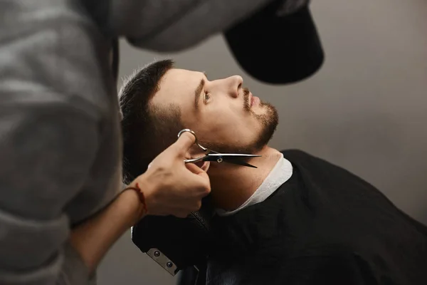 Barber man trims stylish beard of the handsome young brutal man with fashionable haircut who sits in the armchair at a barbershop