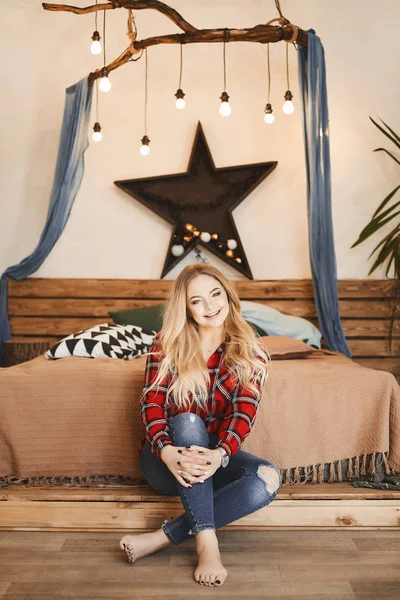 Menina modelo loira feliz e elegante com um sorriso encantador na camisa xadrez vermelha e em jeans senta-se perto da cama de madeira e posando no quarto — Fotografia de Stock