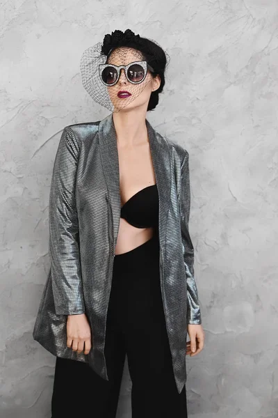 Модная полуголая брюнетка модель девушка с ярким макияжем и в стильной шляпе в расстегнутой серебряной куртке, в черном лифчике и в модных солнцезащитных очках позирует в интерьере — стоковое фото