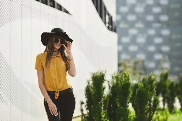 Happy and beautiful brunette model girl with perfect body in the yellow blouse and in the fashionable black hat adjusting her stylish sunglasses, smiles, and posing outdoors — Stock Photo, Image