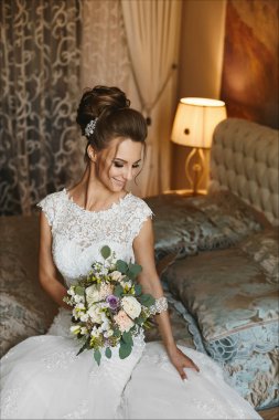 Elleri ve yatak poz yatakta şık bir buket çiçek ile mutlu ve güzel esmer modeli kız parlak makyaj ve beyaz dantel gelinlik çekici gülümseme ile oturuyor