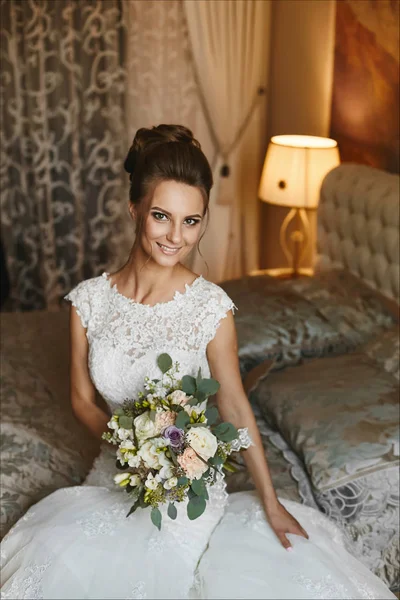 Menina modelo morena feliz e bonita com maquiagem brilhante e sorriso encantador no vestido de noiva de renda branca senta-se na cama com um buquê elegante de flores em suas mãos e posando no quarto — Fotografia de Stock