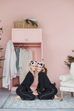 İki güzel, mutlu ve gülümseyen sarışın kız moda pijama ve şık pandalar sit yerdeki şeklinde maskeleri uyku ve iç poz model