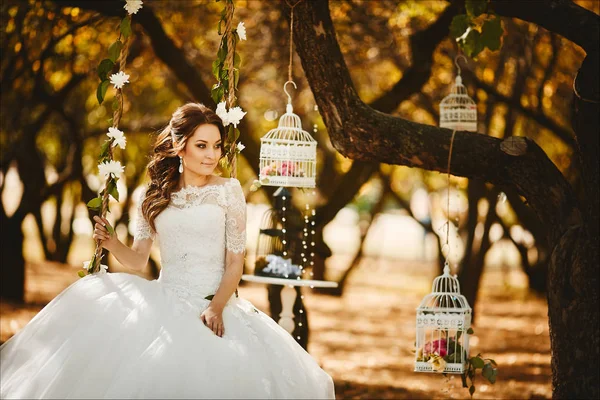 Hermosa y de moda novia joven con peinado de moda en vestido de encaje de boda elegante se sienta en los columpios colgantes, decorado con flores, y posando en el jardín — Foto de Stock