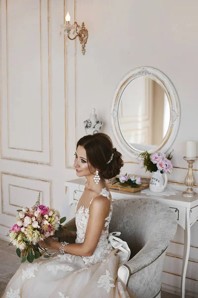 Menina modelo morena bonita e feliz com penteado de casamento elegante em vestido de noiva elegante senta-se na cadeira e posando com um buquê de flores em suas mãos no interior do vintage — Fotografia de Stock