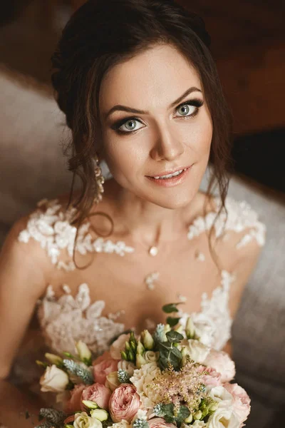 Glücklich brünette Modell Mädchen mit Hochzeitsfrisur und mit schönen Augen in stilvollem Spitzenkleid mit einem Blumenstrauß in ihren Händen posiert im Inneren — Stockfoto