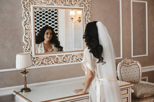 Hermosa y sexy chica modelo morena en peignoir de moda mirando en el espejo en su reflexión en el interior clásico — Foto de Stock