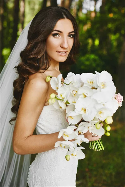 Schöne brünette Modell Mädchen mit hellem Make-up in einem stilvollen Hochzeitskleid mit einem Blumenstrauß in ihren Händen posiert in der Natur — Stockfoto