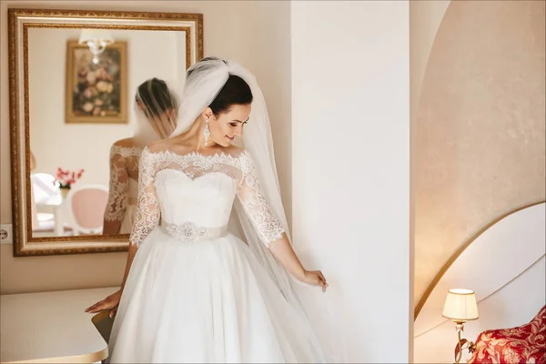 Bella e alla moda bruna in abito da sposa e velo da sposa con grandi orecchini di lusso si erge sullo specchio — Foto Stock