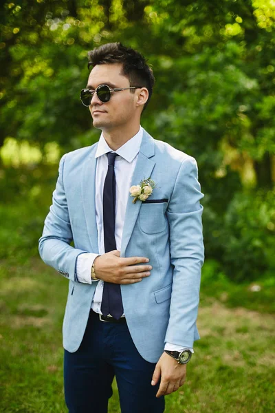 Красивый модель мужчина, одетый в модный синий пиджак с узким галстуком и стильные солнцезащитные очки стоит в зеленом саду — стоковое фото