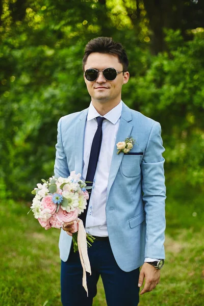Handsome young groom in the fashionable blue jacket with a narrow tie and stylish sunglasses with the bouquet of flowers in his hands