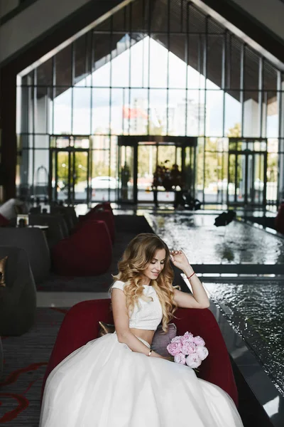Bella ragazza modello bionda con un corpo perfetto in una gonna elegante e camicetta alla moda si siede sulla poltrona con i fiori in mano — Foto Stock