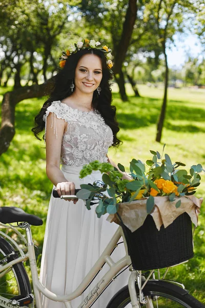 Hermosa chica morena con una corona floral en la cabeza y en un vestido de novia elegante posando con bicicleta en la naturaleza — Foto de Stock