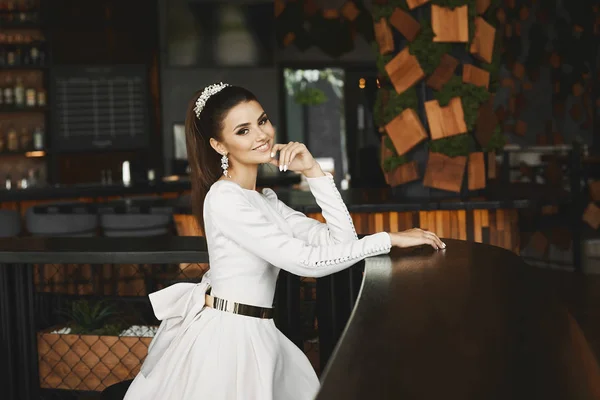 Fiatal elegáns gyönyörű és szexi barna modell nő fehér ruhában, arany öv ül a bárban és a barátja vár — Stock Fotó