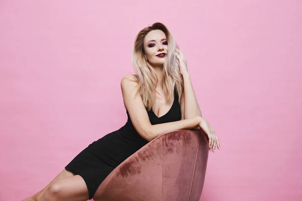 Menina modelo loira sexy e bonita com olhos azuis e maquiagem profissional brilhante em preto elegante vestido apertado senta-se na cadeira, sorrindo e posando no fundo rosa — Fotografia de Stock