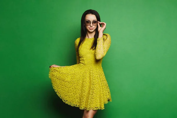 Красивая, сексуальная и модная брюнетка модель девушка в коротком желтом платье и солнцезащитные очки тянет подол ее платье и позирует на зеленом фоне в студии — стоковое фото