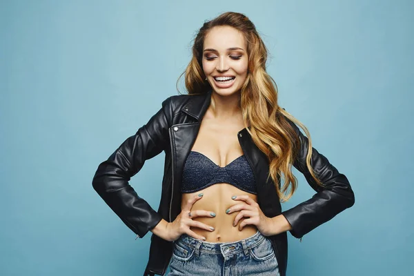 Beautiful and fashionable half-naked blonde model girl with perfect sporty body and professional makeup, in leather jacket, lingerie and jeans smiling and posing with closed eyes at blue background — Stock Photo, Image
