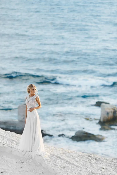 Sensuale e alla moda ragazza modello bionda con corpo perfetto in abito elegante pizzo bianco con schiena nuda, si erge sulla roccia bianca sulla costa del mare — Foto Stock