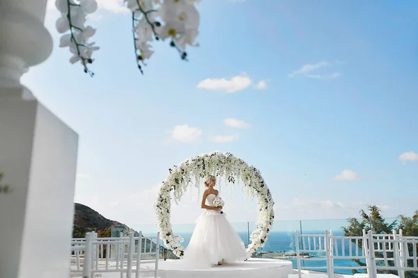 Hermosa chica modelo rubia en el vestido de novia blanco con un ramo de flores en sus manos de pie cerca de arco de boda floral redonda al aire libre —  Fotos de Stock