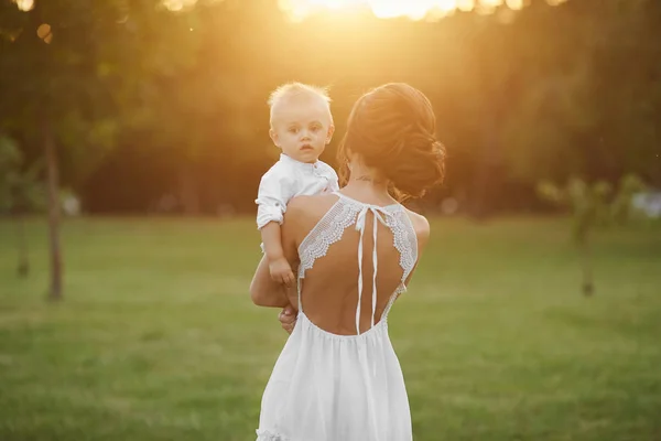 Schöne und sinnliche brünette Modell Mädchen in kurzen weißen Kleid, mit niedlichen kleinen stilvollen Baby Boy auf ihren Händen posiert im grünen Park am frühen Abend bei Sonnenuntergang — Stockfoto