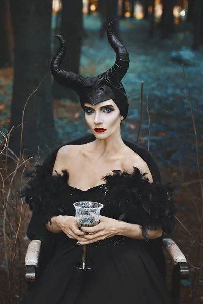 Όμορφο και μοντέρνο μοντέλο slim μελαχρινή κοπέλα στην εικόνα του κακός με ποτήρι κρασί στα χέρια της κάθεται στην πολυθρόνα στο μυστικιστικό δάσος - παραμύθι, cosplay — Φωτογραφία Αρχείου