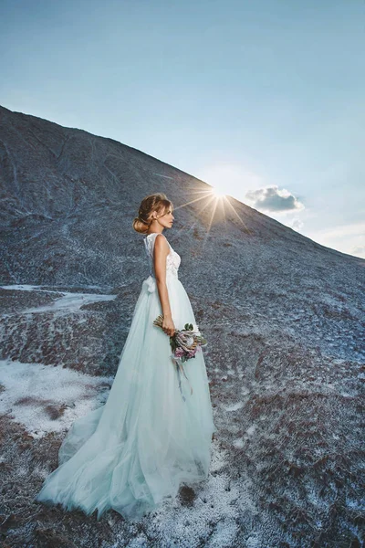 Modische und schöne blonde Model-Mädchen mit Modelfrisur in stilvollem weißen Spitzenkleid mit einem Strauß exotischer Blumen in der Hand posiert auf dem Berg bei Sonnenuntergang — Stockfoto