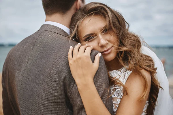 Ragazza modello elegante e felice con acconciatura elegante e con trucco luminoso in un abito di pizzo bianco magra sul suo sposo elegante — Foto Stock