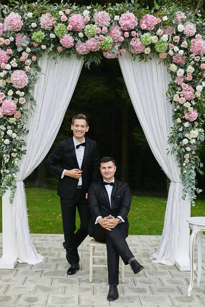 Par av moderiktiga och stiliga unga män i vit skjorta och en snygg svart kostym med fluga, leende och poserar under bröllop arch dekorerad med rosa blommor — Stockfoto