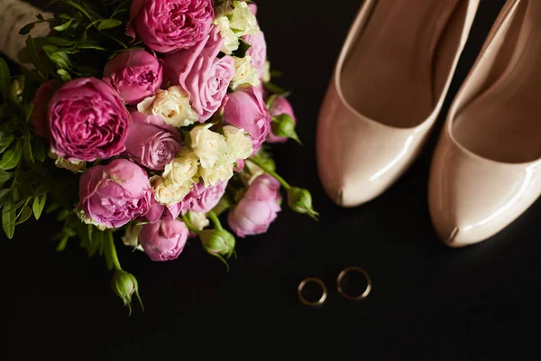 Draufsicht auf Trauringe, Ohrringe, schönen Strauß pinkfarbener Blumen und Frauenschuhe auf schwarzem Hintergrund. Brautmorgen Details Zusammensetzung — Stockfoto